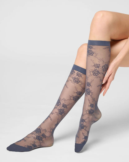 A pair of sheer knee-high lace socks worn on a model's legs, featuring a muted blue-gray floral lace pattern with solid blue-gray cuffs, heels, and toes, set against a light gray studio background.