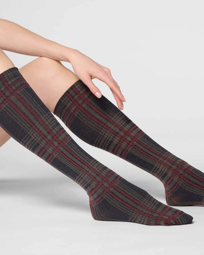 A person wearing knee-high plaid socks in dark gray, black, and red, posed against a light gray studio background.