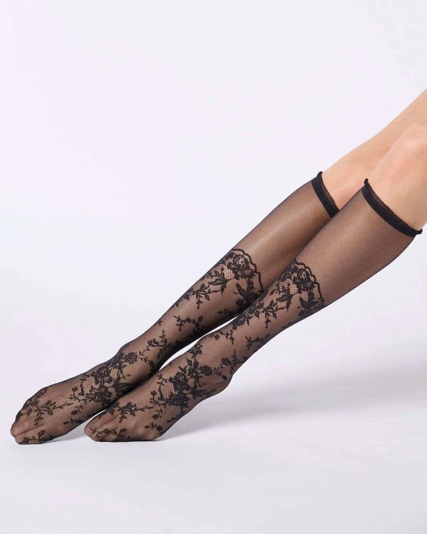 Sheer black knee-high socks with a floral lace pattern from the toe to the calf and a narrow cuff with ruffles.