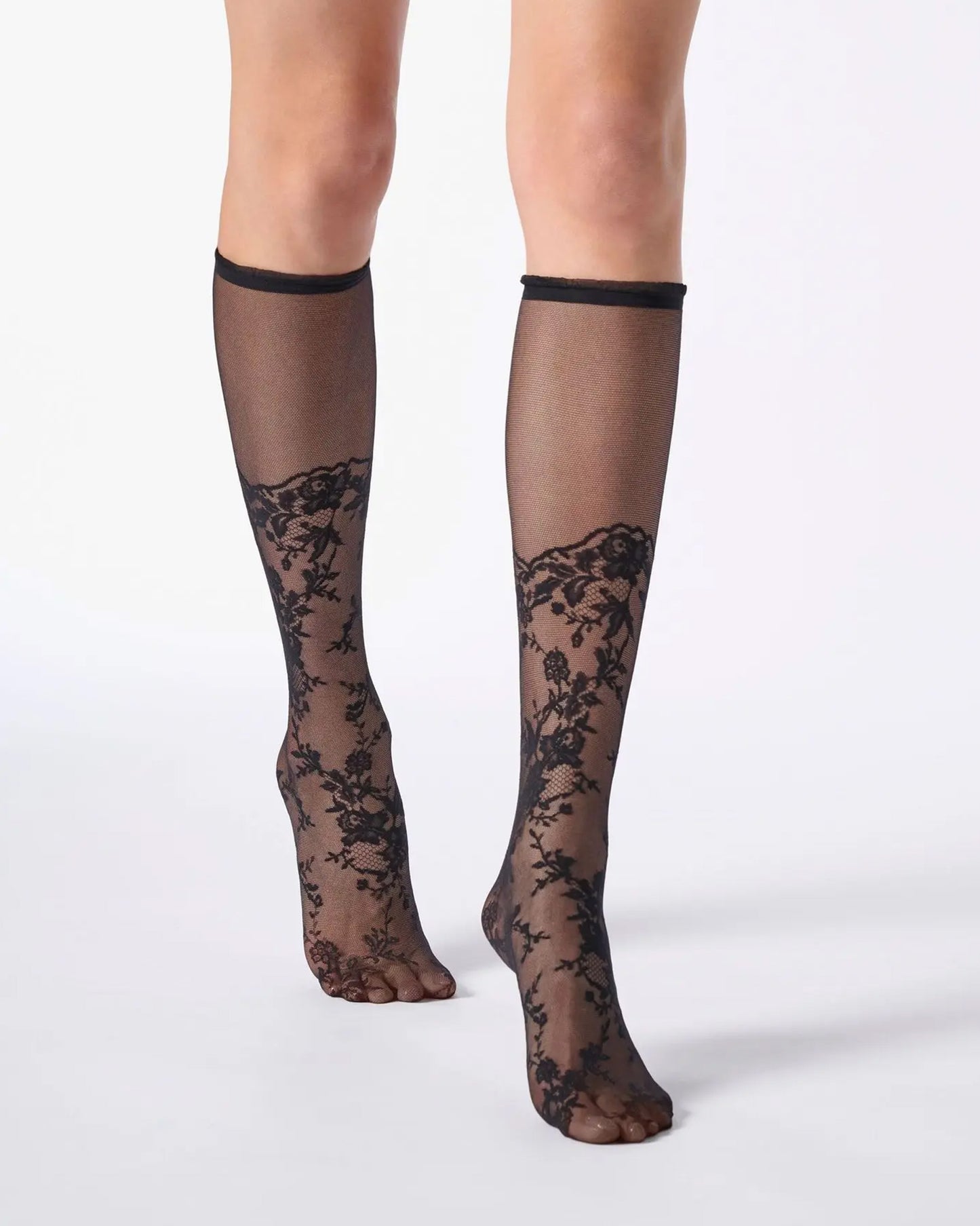Sheer black knee-high socks with a floral lace pattern from the toe to the calf and a narrow cuff with ruffles.