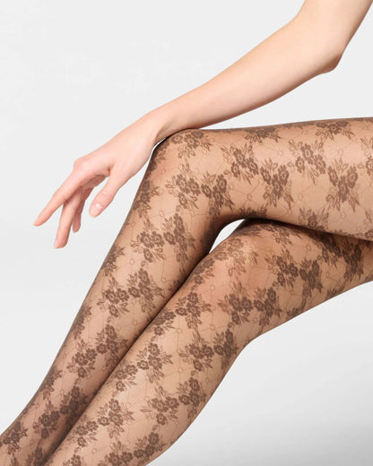 A close-up of a model's legs wearing sheer brown floral lace tights against a light gray studio background.