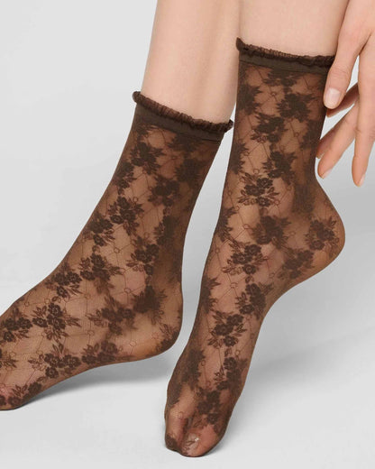 A pair of sheer brown lace ankle socks with a floral pattern and ruffled cuffs, shown on a model's feet against a light gray background.