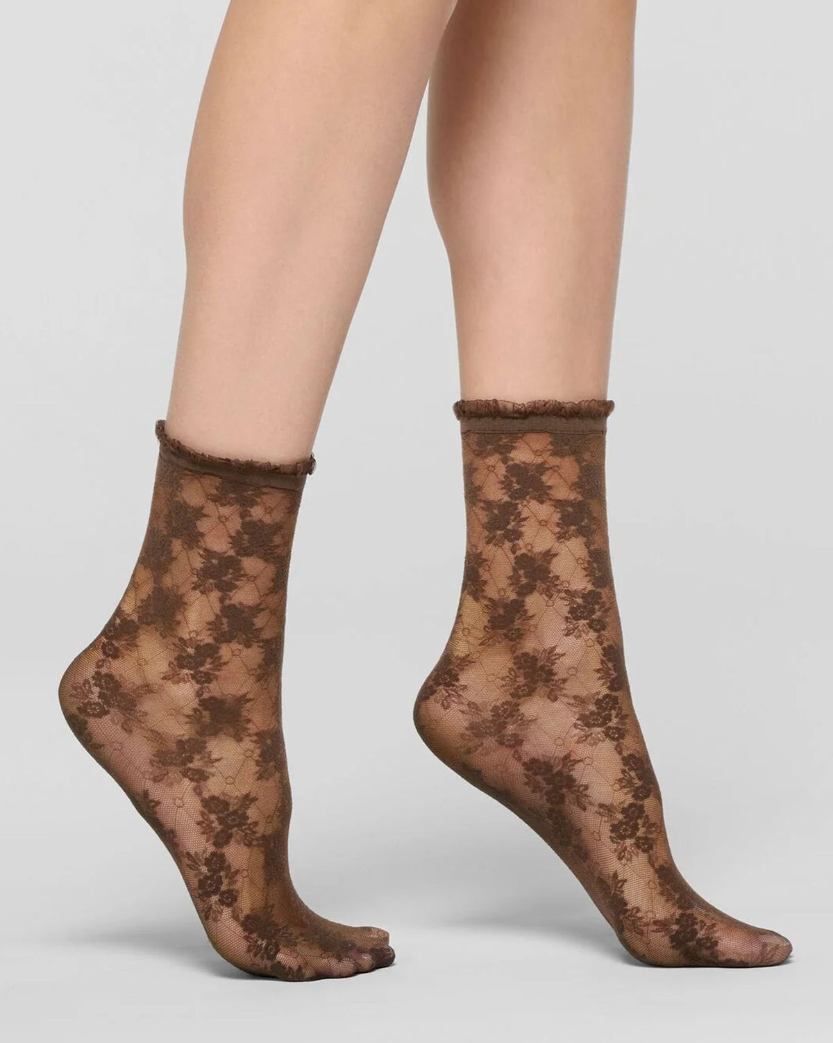A pair of sheer lace ankle socks with a dark brown floral pattern, worn on a model's feet against a light gray studio background.