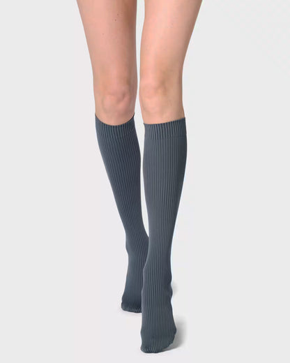 A pair of ribbed knee-high socks in a dark slate gray color, worn by a model against a light gray studio background.