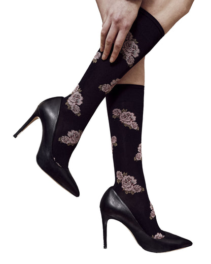 A person wearing black knee-high opaque socks with a muted floral pattern, paired with black pointed-toe stiletto heels, shown against a plain white background.