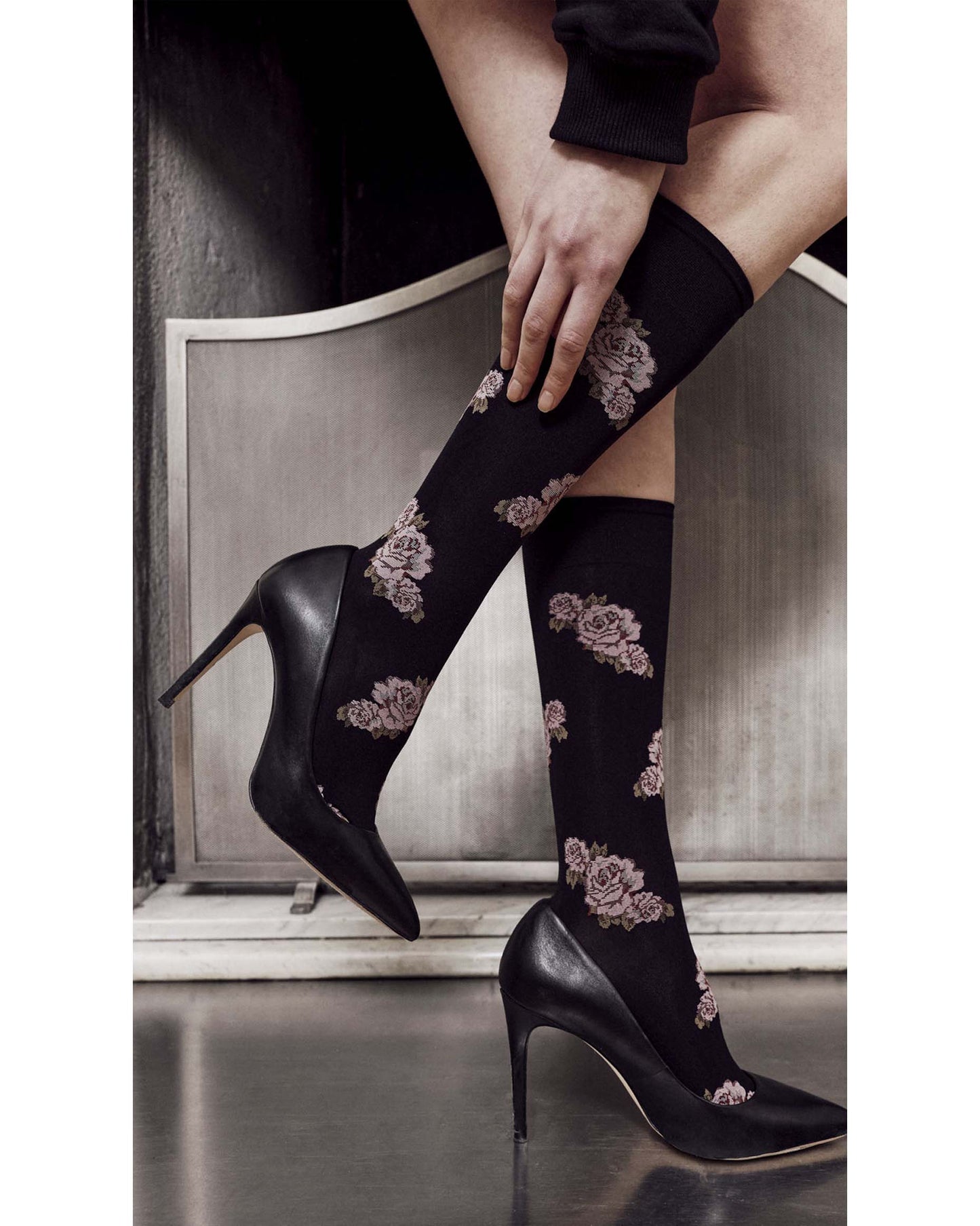 A person wearing black knee-high socks with a dusty rose floral pattern, paired with black pointed-toe stiletto heels, posed in front of a gray metal panel.