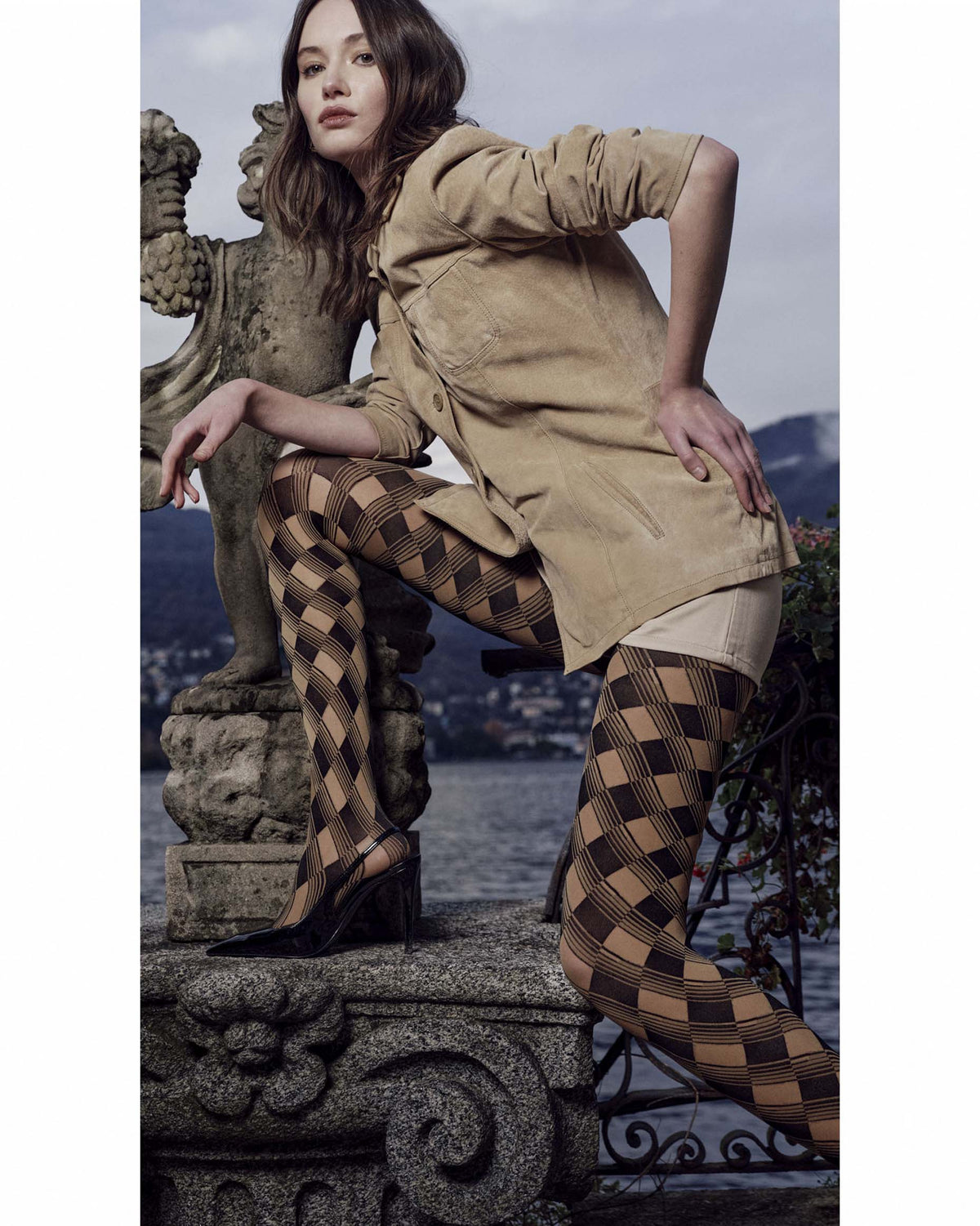 A model wearing black and beige argyle hosiery with a tan shirt and beige shorts, posing on a stone ledge with a scenic lake and mountains in the background.