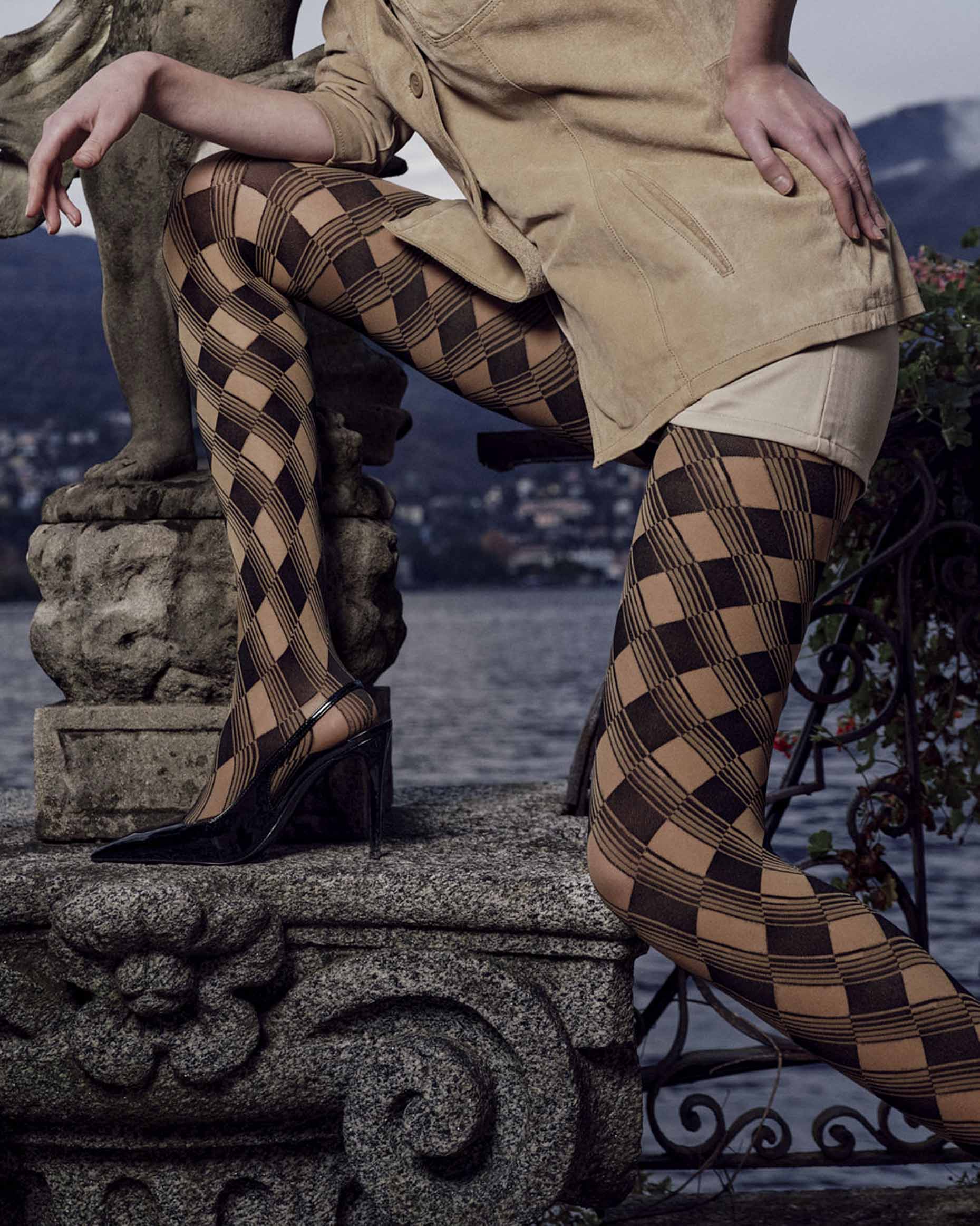 A model wearing black and camel checkered harlequin tights, styled with a tan trench coat and black high heels, posing on a stone fountain next to a lake with mountains in the background.