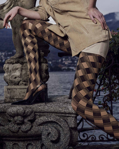 A model wearing black and camel checkered harlequin tights, styled with a tan trench coat and black high heels, posing on a stone fountain next to a lake with mountains in the background.