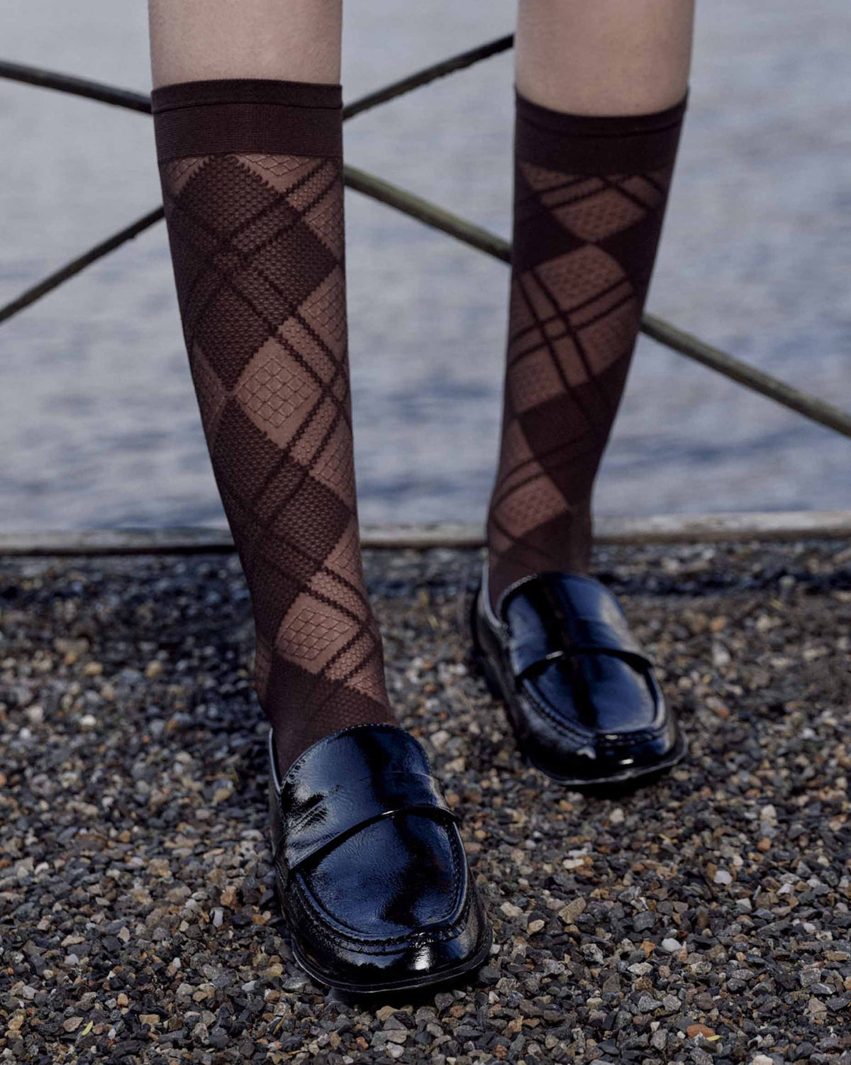 A person wearing dark brown argyle patterned knee-high socks and shiny black leather loafers, standing on a gravel surface near water.