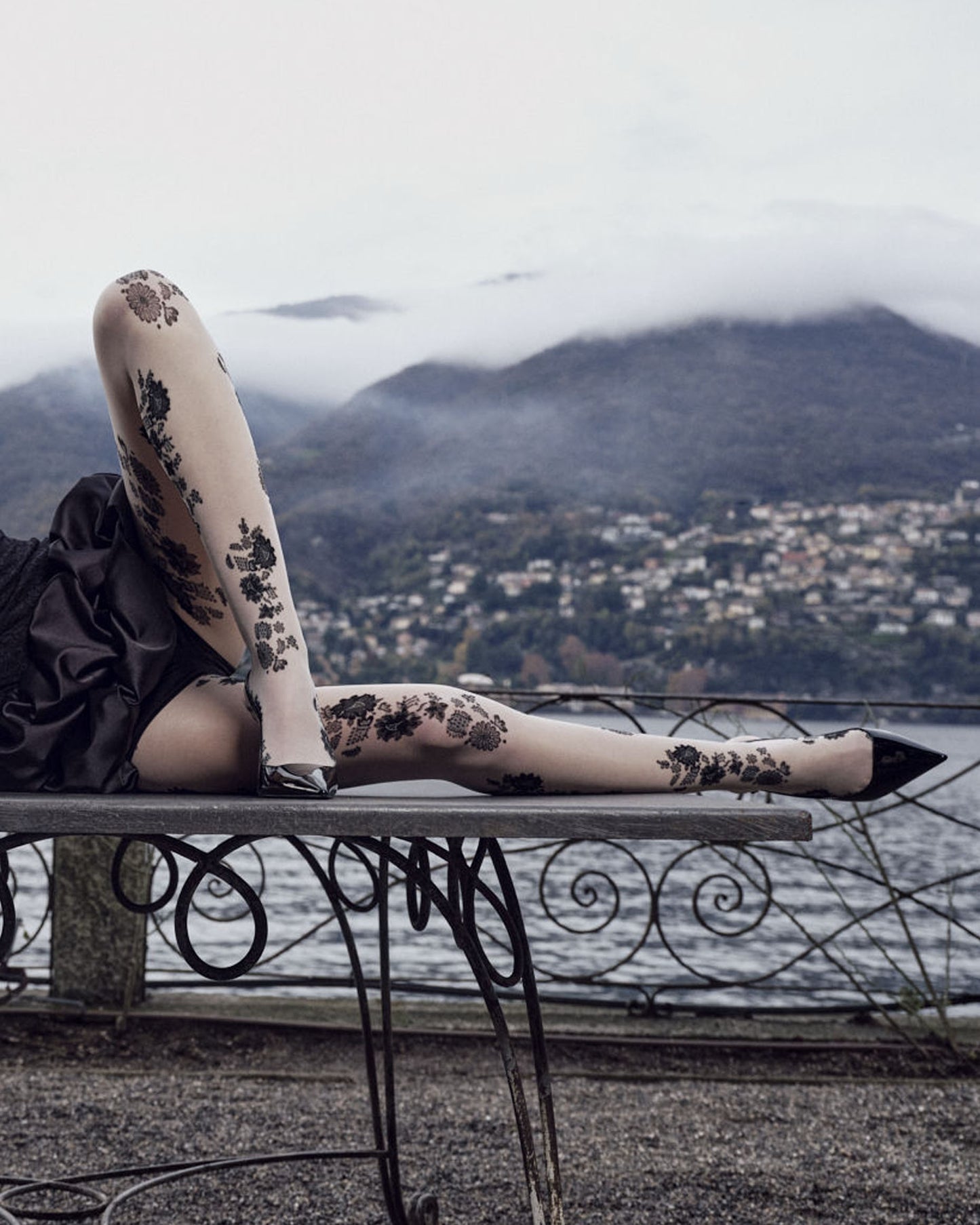 Person wearing floral-patterned tights sitting on a metal bench with a scenic background of mountains and water.