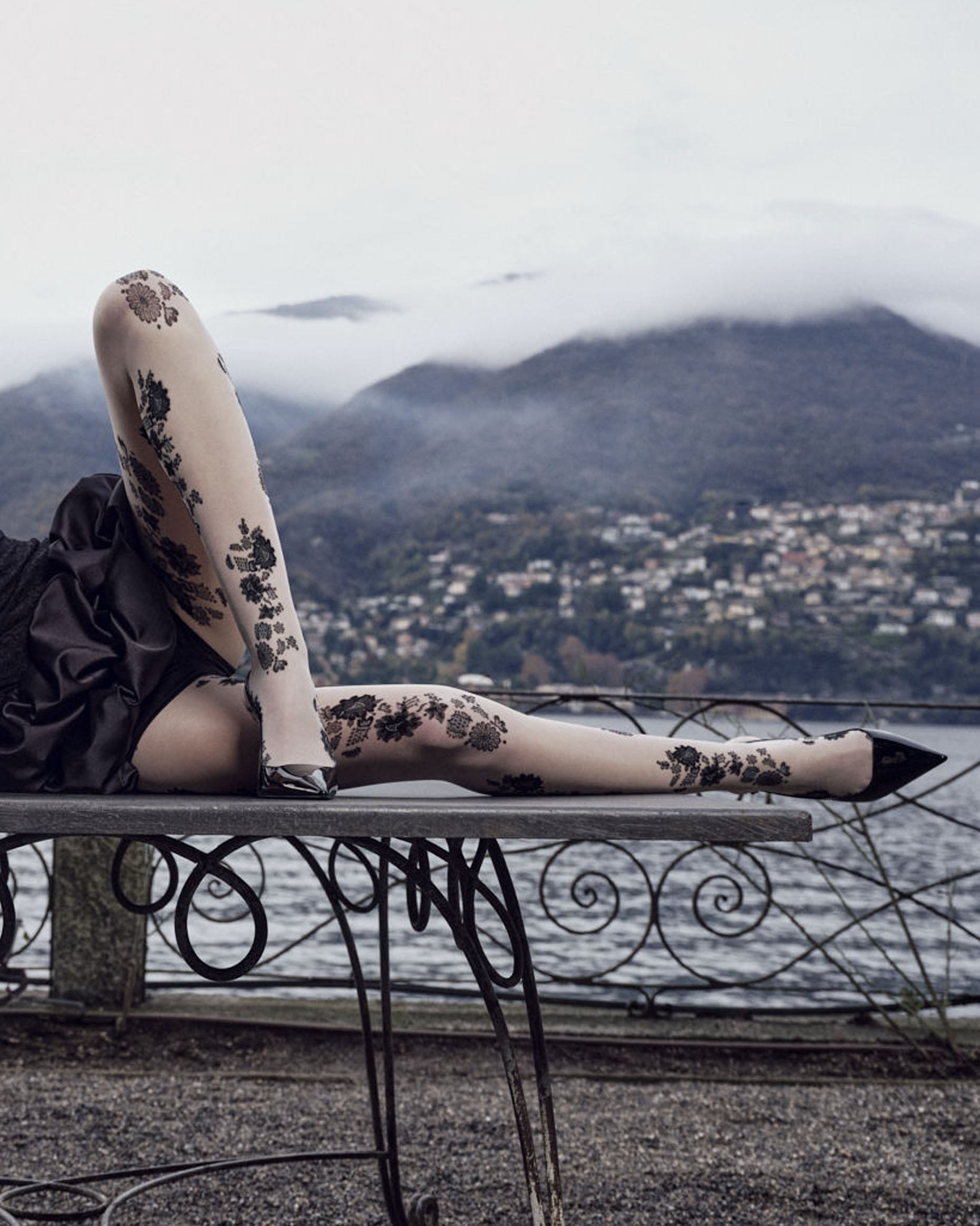 Person wearing floral-patterned tights sitting on a metal bench with a scenic background of mountains and water.