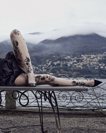 Person wearing floral-patterned tights sitting on a metal bench with a scenic background of mountains and water.