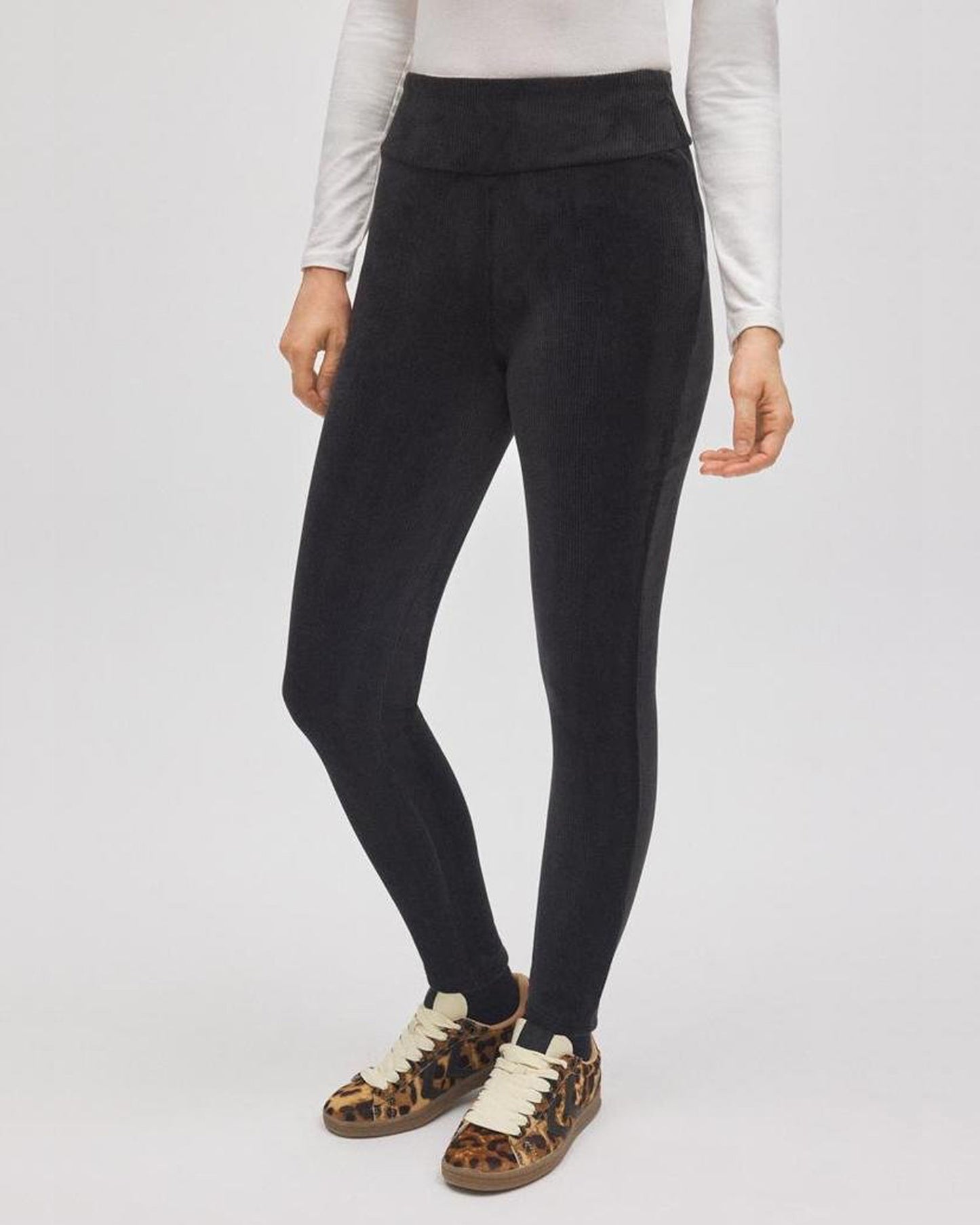 Person wearing black velvet leggings with a white top and brown shoes on a light gray background
