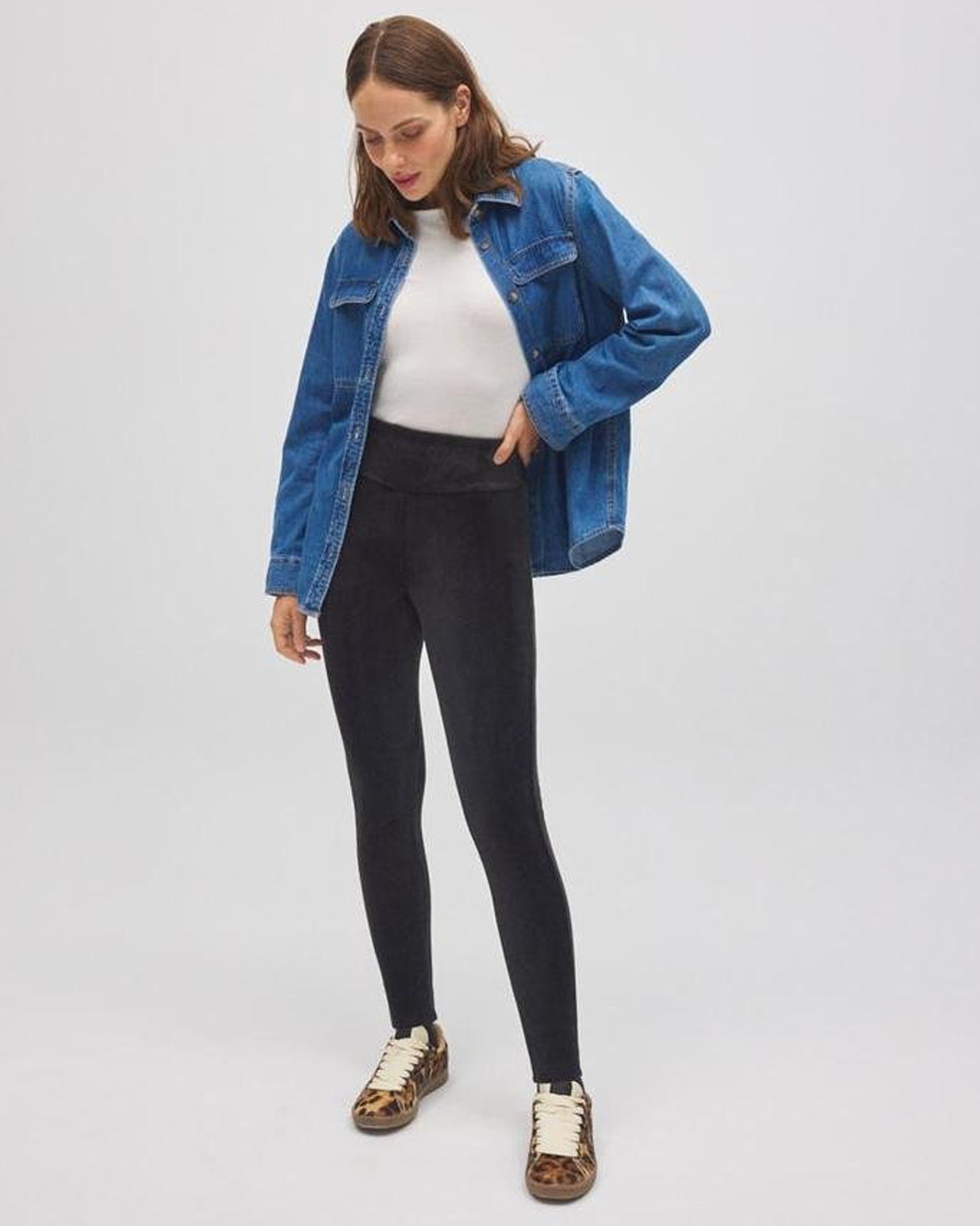 Woman wearing a blue denim jacket, white top, black leggings, and patterned shoes on a light gray background